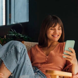 Eine Person sitzt in einem Sessel und blickt lachend auf ihr Smartphone, das sie in der Hand hält.