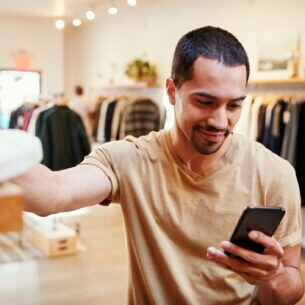 Eine Person steht in einem Bekleidungsgeschäft, greift mit einer Hand nach einem Paar Sneaker und hält in der anderen Hand ein Smartphone.