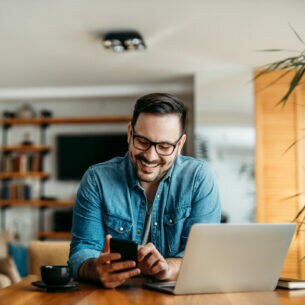 Eine Person sitzt an einem Tisch und blickt auf ein Smartphone, das sie in den Händen hält. Vor ihr stehen ein geöffnetes Laptop und eine Kaffeetasse.