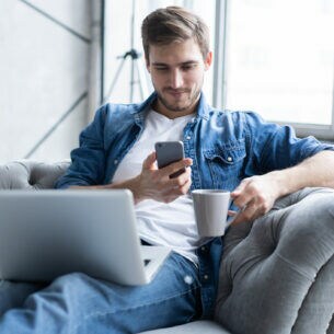 Eine Person sitzt mit ausgestreckten Beinen auf einem Sofa, hat auf dem Schoß ein geöffnetes Laptop liegen und hält in ein Smartphone sowie eine Tasse in den Händen. 