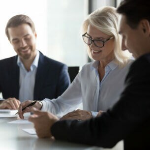 Drei Personen in Businesskleidung sitzen an einem Tisch, auf dem sich ein aufgeklappter Laptop und Papierunterlagen befinden.