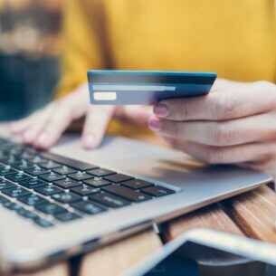 Nahaufnahme einer Person vor einem geöffneten Laptop, in einer Hand eine Bankkarte haltend, die andere liegt auf der Laptoptastatur.
