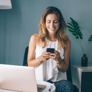 Influencerin sitzt mit Smartphone und Laptop in ihrem Wohnzimmer.