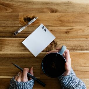 Leere To-do-Liste auf einem Holztisch mit Kaffeetasse und Stift in den Händen einer Person.