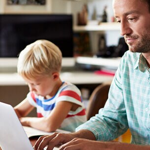 Blogbild: Person arbeitet am laptop während ein Kind daneben Hausaufgaben macht