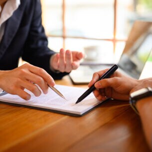 Artikelbild Eine Person im Anzug zeigt auf Dokumente und eine weitere Person unterzeichnet die Dokumente