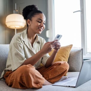 : Eine Frau sitzt im Schneidersitz auf einer Couch und schaut lächelnd auf ihr Smartphone. Vor ihr steht ein Laptop.