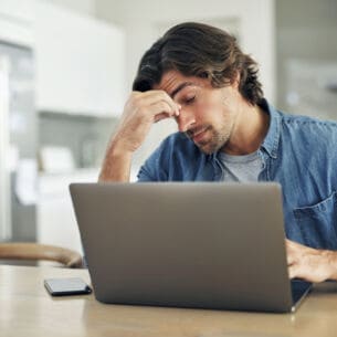 Ein Mann sitzt vor einem Laptop an einem Tisch, hat die Augen geschlossen und eine Hand an seine Stirn gelegt.