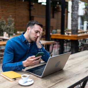 Ein Mann sitzt an einem Tisch vor einem Laptop und hält ein Smartphone in der Hand. Er hat die Augen geschlossen und legt eine Hand über sein Gesicht.