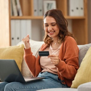 Eine Person sitzt lächelnd auf einem Sofa, einen Laptop auf den Beinen und eine Kreditkarte in der Hand und ballt eine Faust.