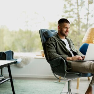 Eine Person sitzt vor einer großen Fensterfront in einem Sessel mit einem Laptop auf dem Schoß.