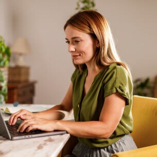 Eine Frau sitzt an einem Tisch vor einem Laptop und legt ihre Hände auf die Tastatur.