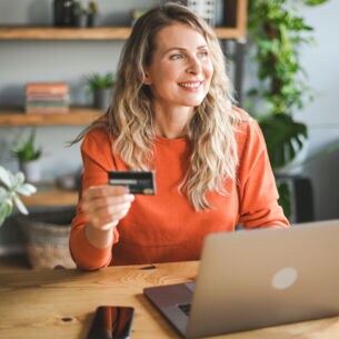 Eine Frau sitzt lächelnd an einem Tisch. Vor ihr steht ein Laptop und in der Hand hält sie eine Kreditkarte.