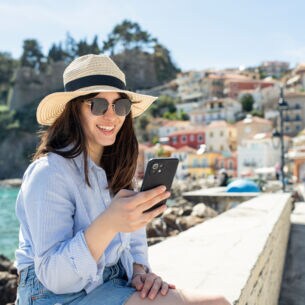Person schaut auf ihr Handy während sie auf einer Mauer sitzt. Hinter ihr ist das Meer und bunte Häuser