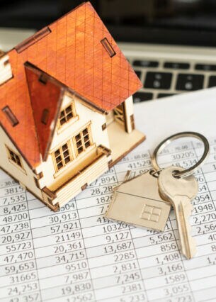 Ein kleines Hausmodell aus Holz steht auf einem Blatt Papier mit vielen Zahlen, daneben ein Schlüsselbund