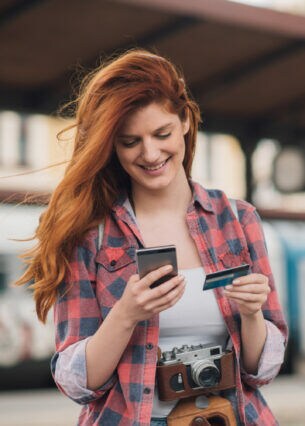 Eine lächelnde junge Frau am Bahngleis mit Smartphone in der einen, Kreditkarte in der anderen Hand