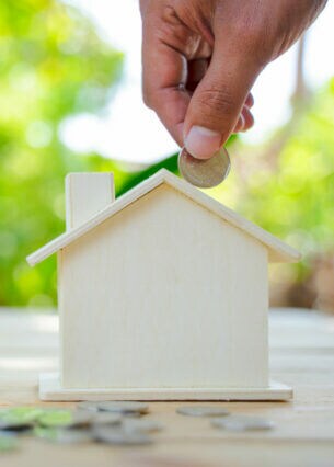 Nahaufnahme einer Hand, die Münzen in eine Spardose in der Form eines Hauses steckt