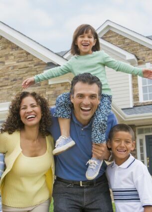 Eine Frau, ein Mann und drei Kinder, eins davon auf den Schultern des Mannes, lachen in die Kamera. Im Hintergrund steht ein Einfamilienhaus.