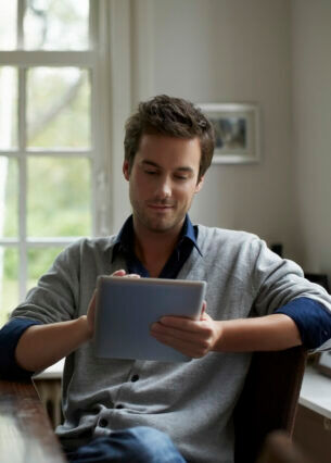 Mann sitzt an einem Tisch und schaut lächelnd auf ein Tablet in seiner Hand.