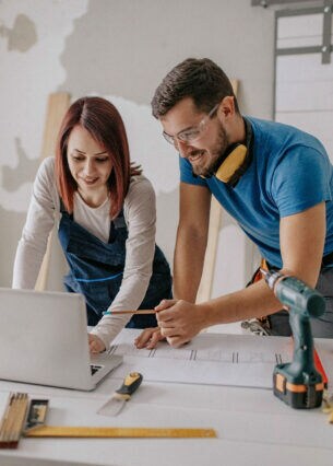Ein Mann und eine Frau in Arbeitskleidung stehen in einem renovierungsbedürftigen Zimmer und blicken auf ein Laptop.