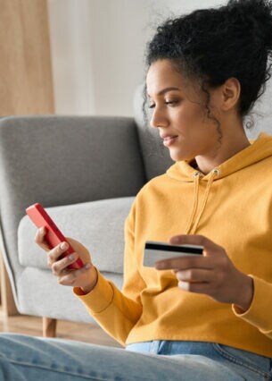 Junge Frau mit gelbem Pullover an ein Sofa gelehnt mit Kreditkarte in der einen und Smartphone in der anderen Hand