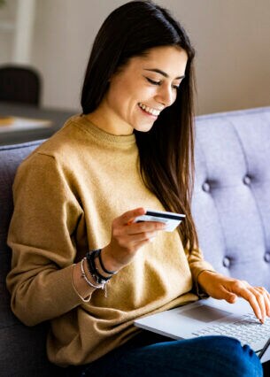 Eine Frau im Schneidersitz auf einem Sofa mit Laptop im Schoß und Kreditkarte in der Hand