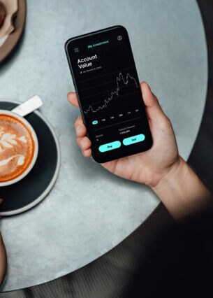 Blickwinkel einer Person, die beim Kaffeetrinken einen Aktienkurs auf ihrem Smartphone überprüft