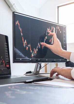 Eine Person, deren Arme zu sehen sind, an einem Schreibtisch mit zwei Computern, auf deren Bildschirmen Kursverläufe zu sehen sind