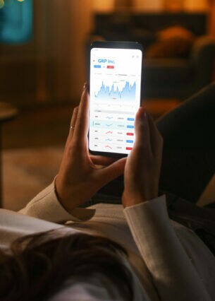 Eine Frau liegt auf dem Sofa und blickt auf ihr Smartphone mit Aktienkursten, das sie in ihren Händen hält.