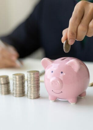 Eine Person am Tisch, die mit einer Hand in ein Heft schreibt und mit der anderen eine Münze in ein Sparschwein steckt, neben dem Münzstapel platziert sind