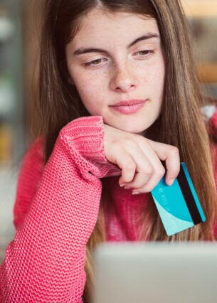 Ein Mädchen im Teenageralter sitzt in einem Café vor einem Laptop und hält eine Bankkarte in der Hand.