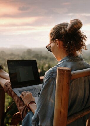 Eine Frau sitzt im Freien auf einer Terrasse, arbeitet an einem Laptop mit Blick ins Grüne.