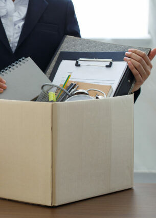 Eine Person im Anzug steht vor einem gepackten Karton, der auf einem Schreibtisch steht.
