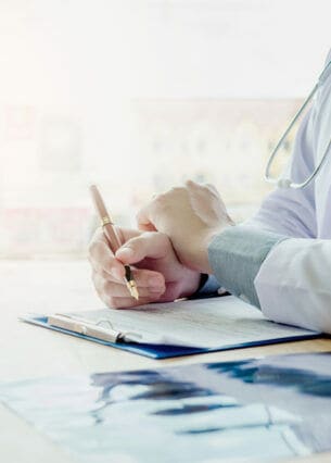 Zwei Personen sitzen sich gegenüber an einem Tisch, eine davon trägt einen Arztkittel und schreibt etwas auf ein Dokument