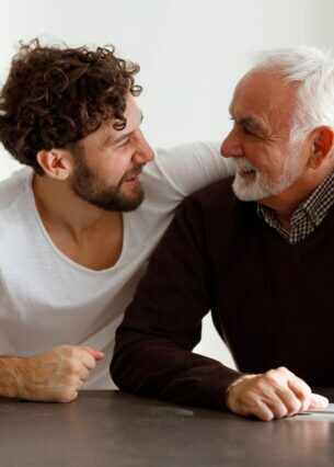 Ein lachender Mann mit grauem Bart und grauen Haaren wird von einem lachenden anderen Mann umarmt.