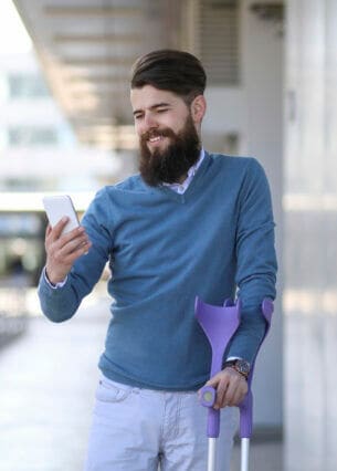 Mann mit Krücken steht vor einem Gebäude und blickt lächelnd auf sein Smartphone.
