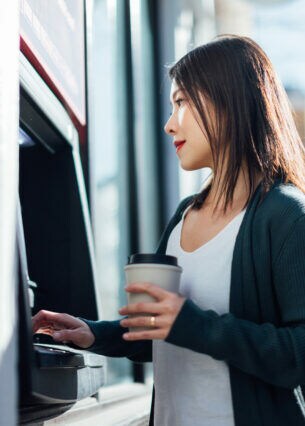 Eine junge Frau mit einem Becher in der einen Hand in einer Straße vor einem Geldautomaten