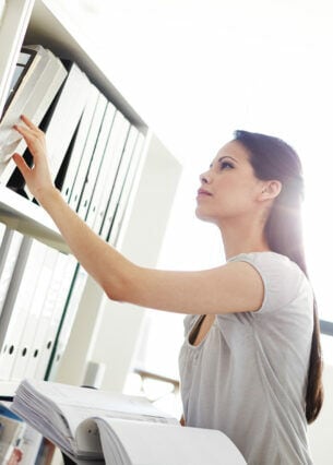 Eine Frau hält einen aufgeschlagenen Aktenordner in der Hand und durchsucht ein Regal mit weiteren Ordnern