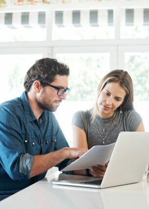 Zwei Personen sitzen an einem Tisch vor einem Laptop und betrachten ein Dokument