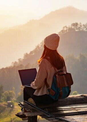 Eine Person mit Laptop sitzt auf einem hölzernen Untergrund und blickt auf ein Naturpanorama mit Bergen und Sonnenaufgang