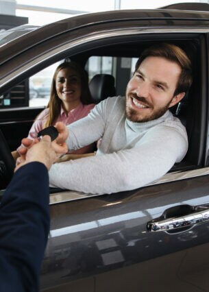 Eine Frau in einem Auto neben einem Mann, der von einer Person durch das Autofenster einen Autoschlüssel annimmt