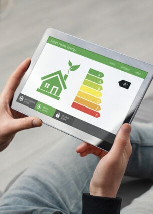Blick über die Schulter einer Person, die ein Tablet in den Händen hält, auf dem Energieeffizienzklassen und de schematische Darstellung eines Hauses abgebildet sind.