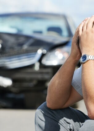 Eine Person hockt am Straßenrand und hat den Kopf in die Hände gestützt, im Hintergrund steht ein kaputtes Auto