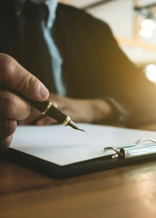 Eine Person am Tisch mit einem Füllfederhalter in der Hand über ein Papier mit Klemmbrett, neben dem ein Richterhammer liegt