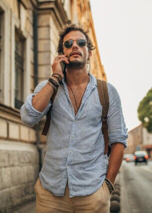 Mann mit Bart, Sonnenbrille und Rucksack auf dem Rücken steht auf einem Bürgersteig und telefoniert