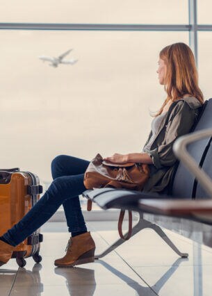 Eine Person sitzt mit einem Koffer im Flughafen und blickt aus dem Fenster, an dem gerade ein Flugzeug vorbeifliegt