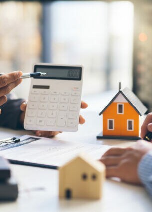 Zwei Personen sitzen am Tisch, die eine unterschreibt ein Dokument, die andere hält einen Taschenrechner in der Hand und zeigt mit einem Stift darauf.