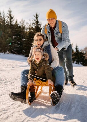 Eine dreiköpfige Familie im Schnee, von der zwei Personen auf einem Schlitten sitzen und die dritte Person anschiebt.
