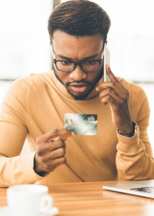Eine Person sitzt telefonierend vor einem geöffneten Laptop am Tisch und blickt auf seine Bankkarte in der Hand.