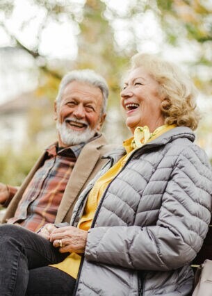 Zwei ältere Menschen sitzen lachend auf einer Parkbank.